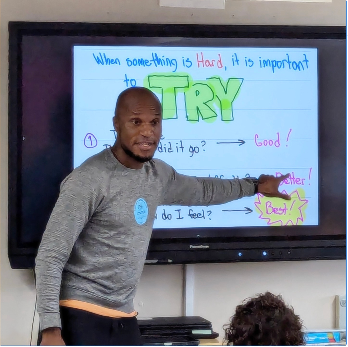 A man points to a whiteboard with colorful text saying, 'When something is hard, it is important to TRY,' highlighting steps on progress and feelings like good, better, and best.