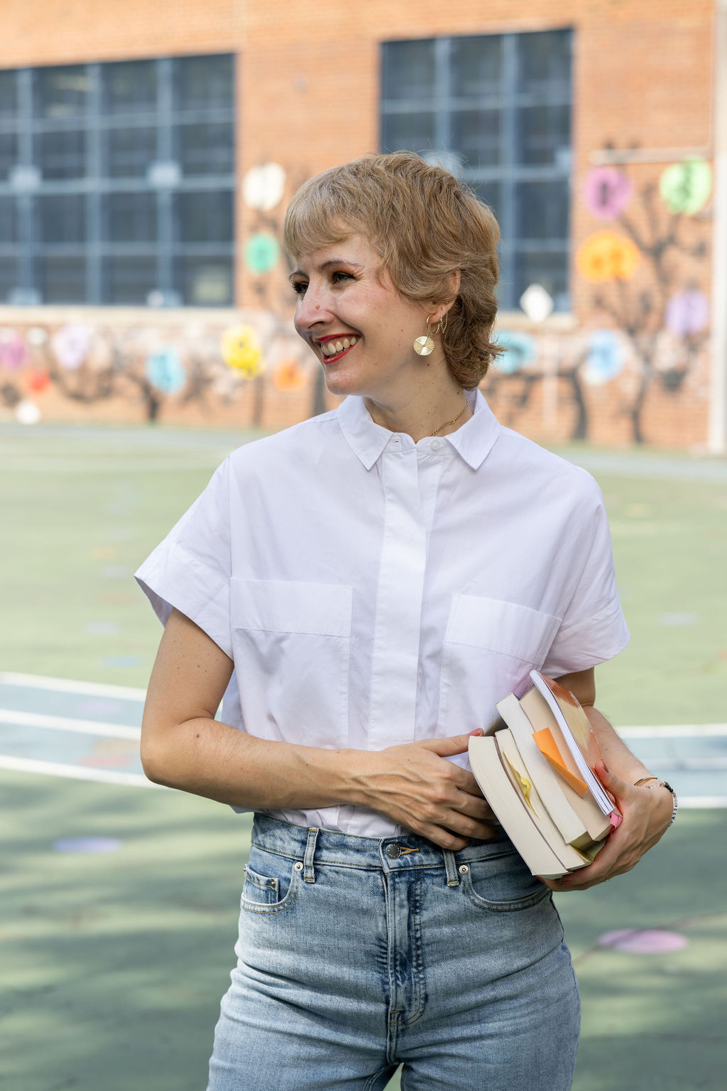 Smiling woman wearing a white shirt and jeans holding a stack of books outdoors with a colorful mural in the background.