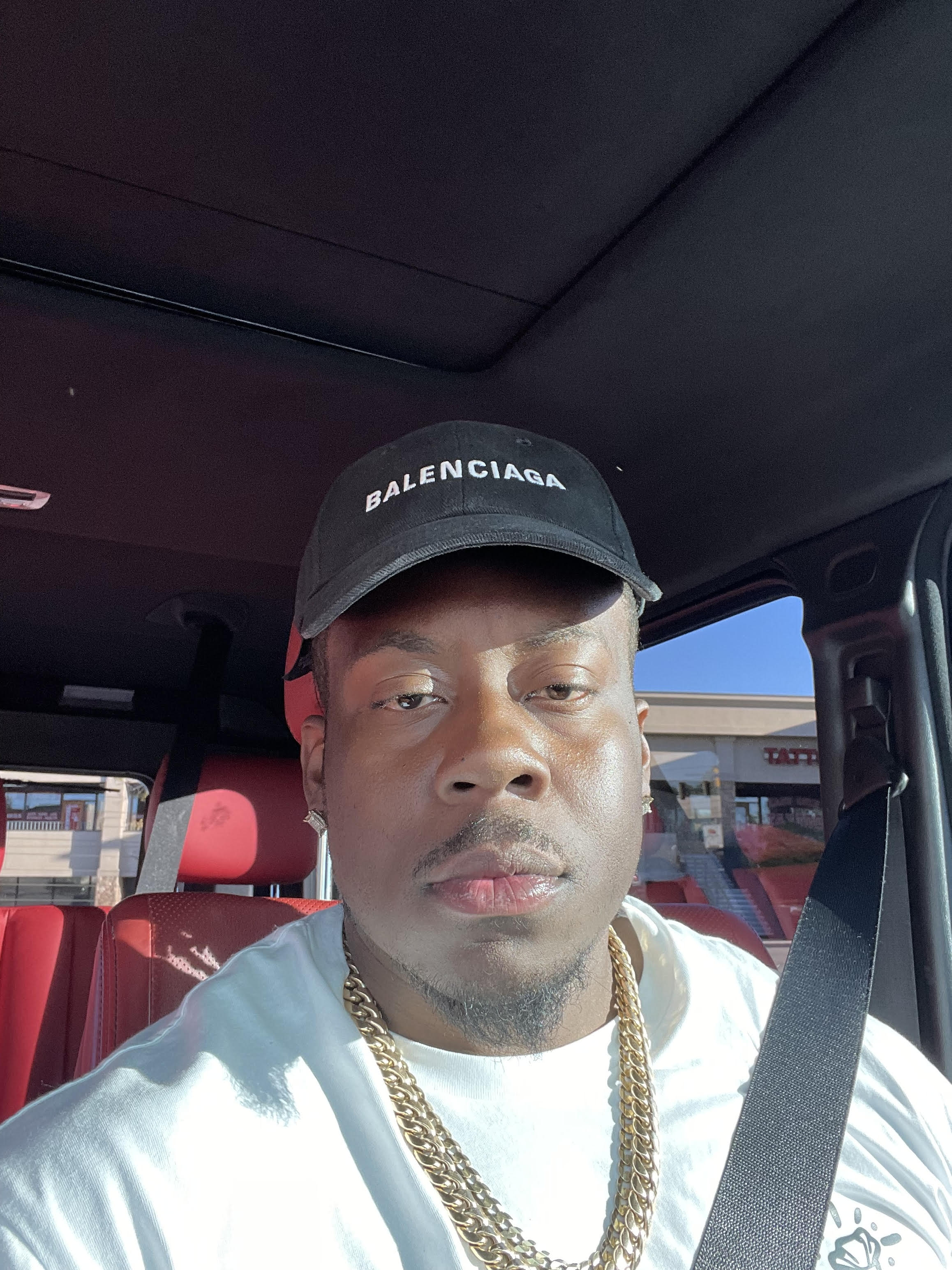 Man wearing a black Balenciaga cap, gold chains, and white shirt sitting in a car with red seats and a seatbelt across his chest.