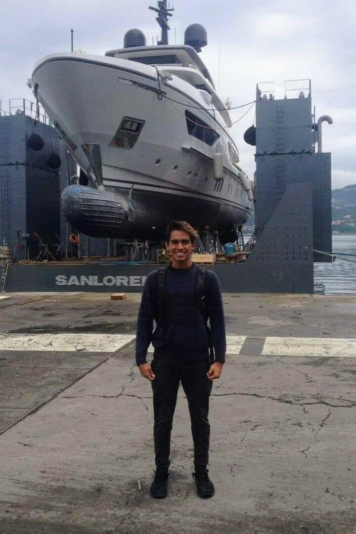 Cristo León standing in front of a large yacht at a shipyard