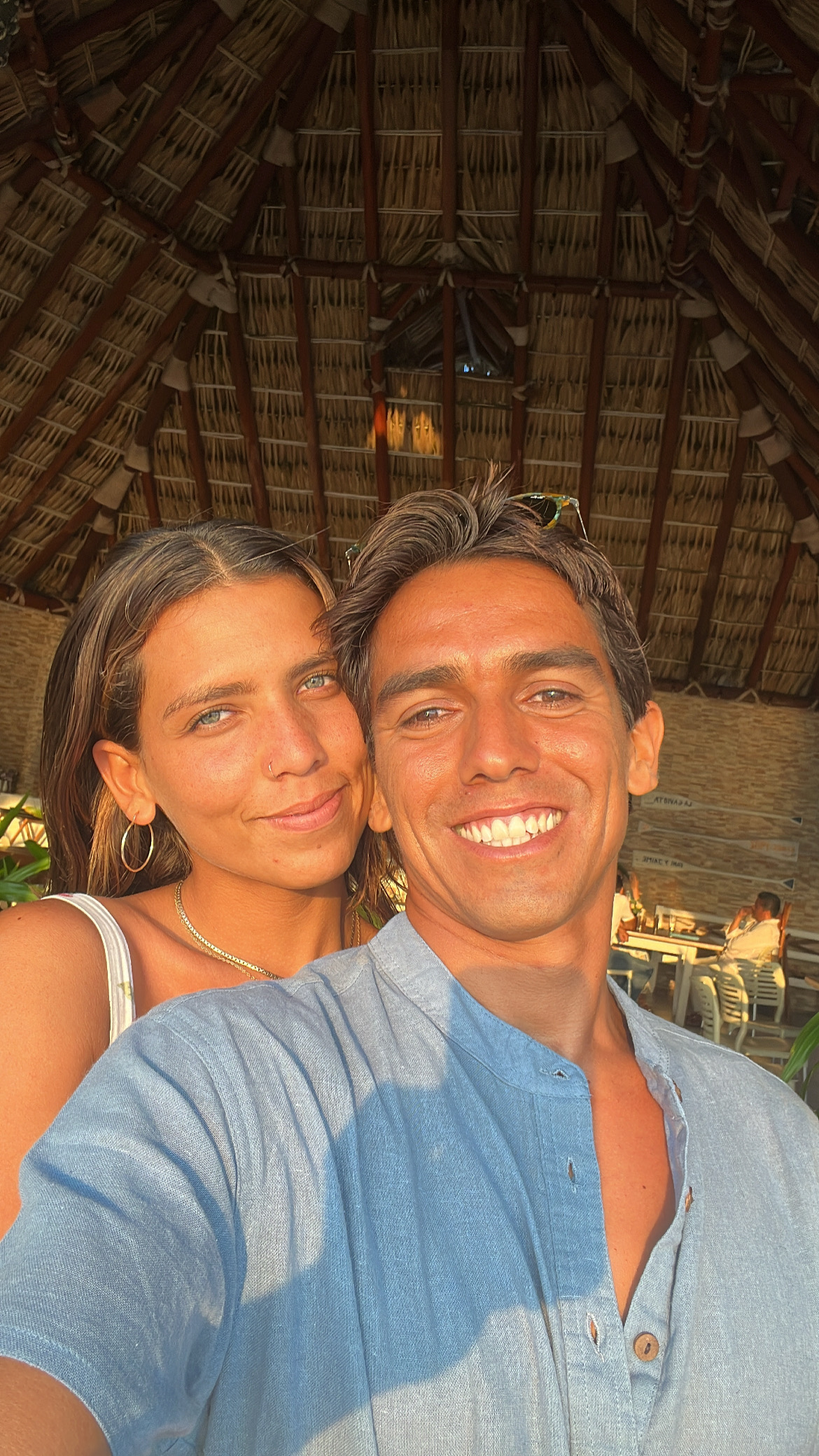 Cristo León and Brigit smiling together under a palm-thatched roof at sunset