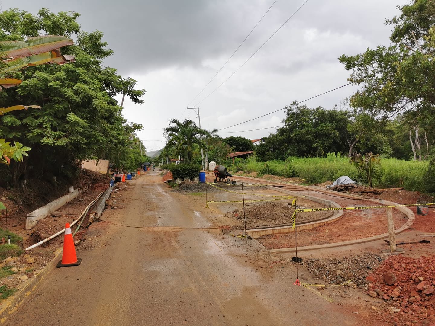New curbs and sidewalk forms under construction beside a dirt road, with a cement mixer and caution tape