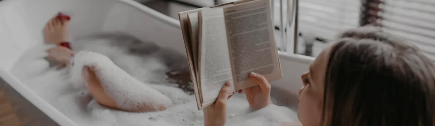 Person in a bubble bath reading a book with legs resting on the tub edge.