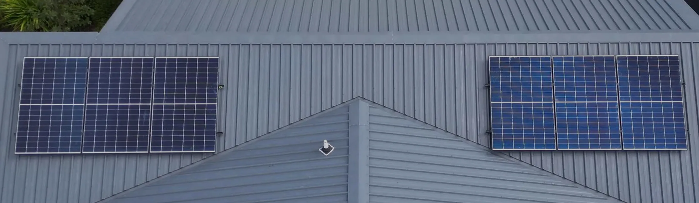 Six solar panels installed on a slanted gray metal roof of a house, three on each side of a central peak.