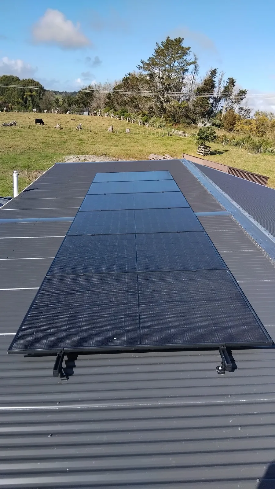 Black solar panels installed on a gray metal roof with green fields and trees in the background.