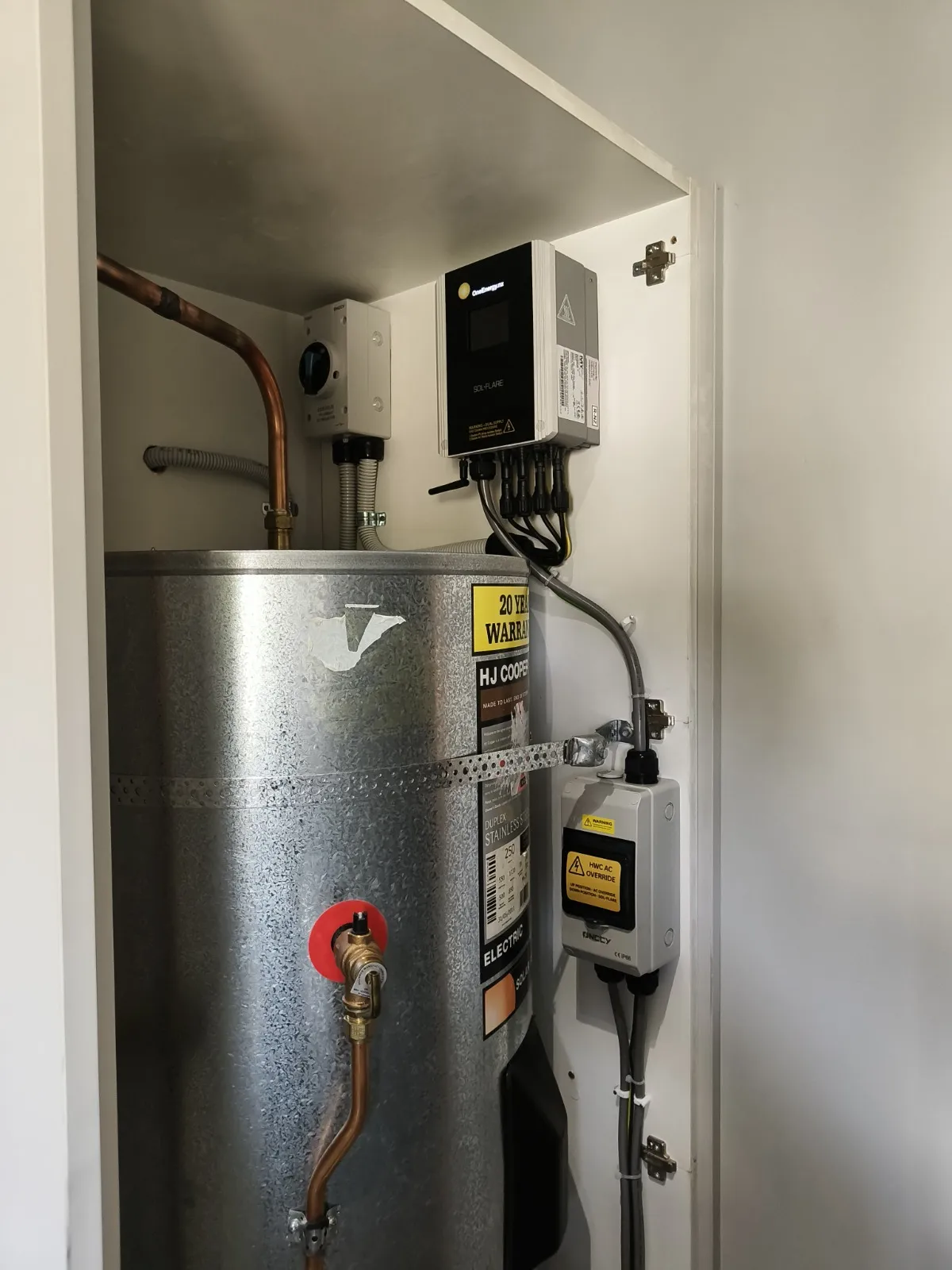 Electric water heater tank with copper pipes and mounted control boxes in a white cabinet.