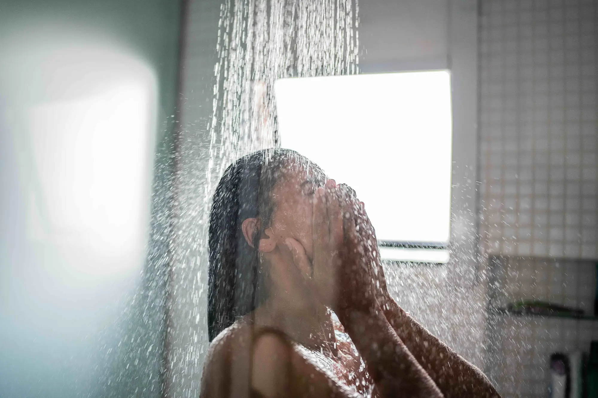Person washing their face under a running shower in a bathroom.