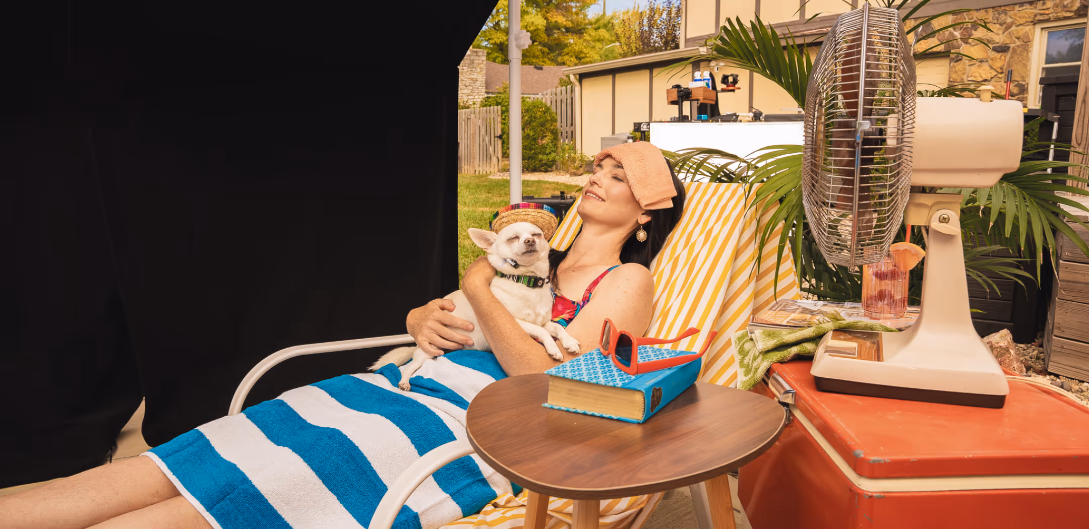 Woman relaxing on a striped lounge chair with a small dog wearing a colorful hat, a striped towel, a blue book with red sunglasses on a wooden side table, and an electric fan nearby in a backyard setting.
