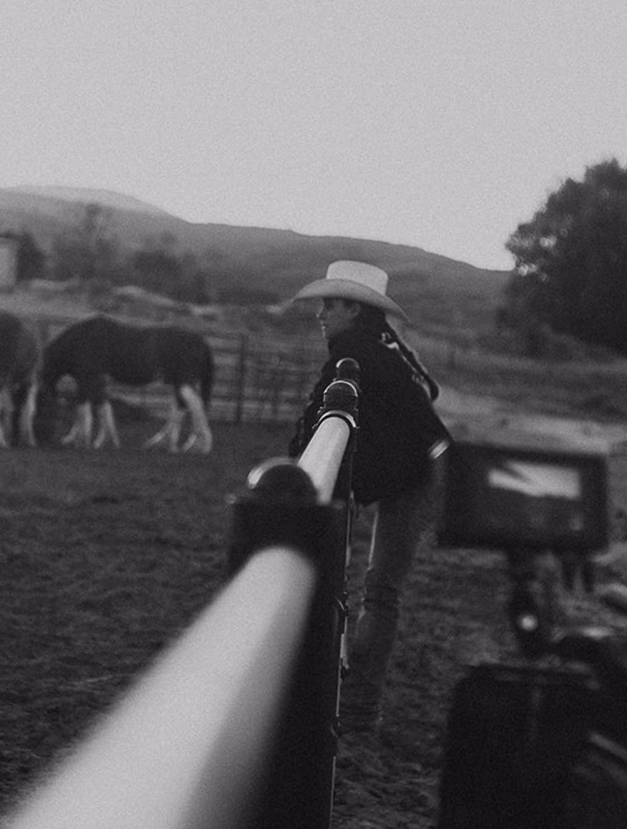 Black and white photo of a person in a cowboy hat leaning on a fence with horses grazing in the background.