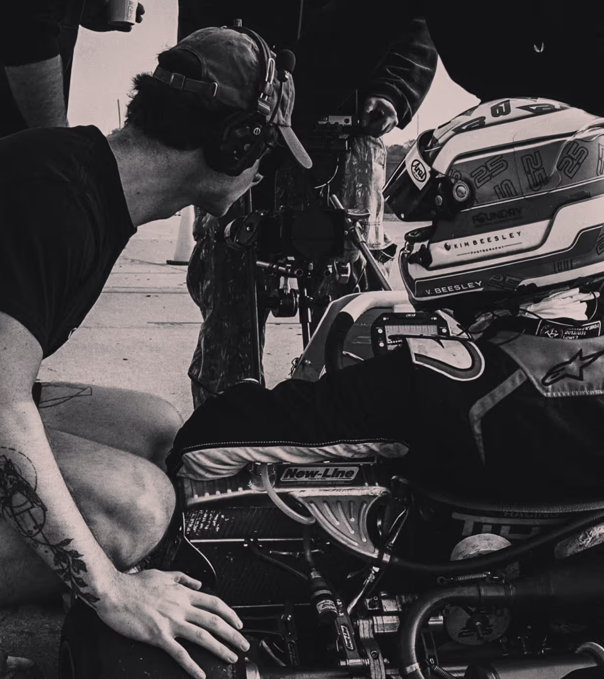 Man wearing headphones and a cap crouches beside a go-kart racer in helmet and suit, inspecting the vehicle.