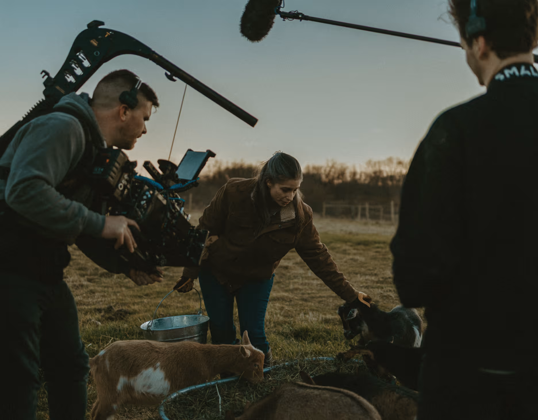A woman feeding and petting goats in an outdoor field while a camera operator films her with professional equipment.