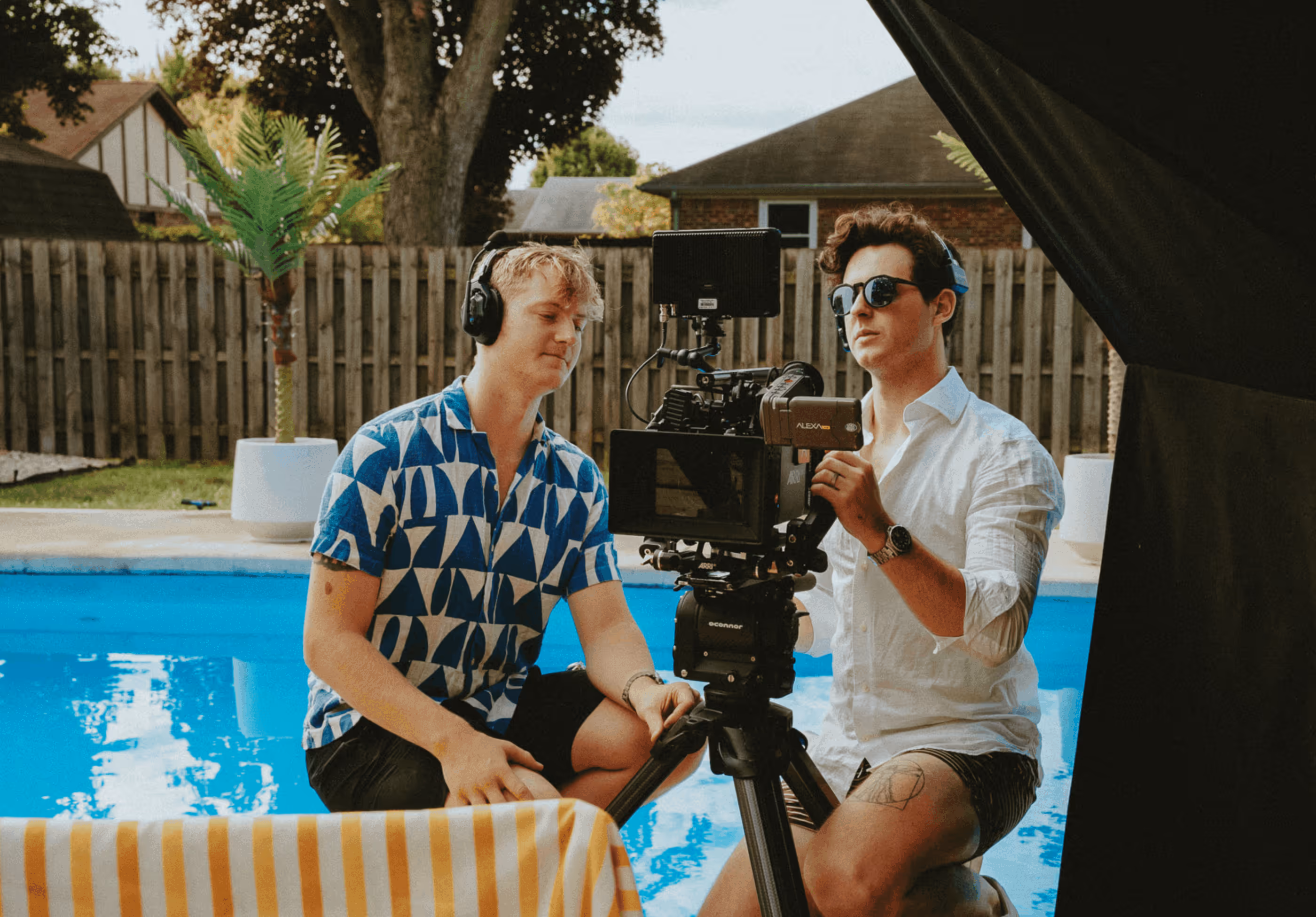 Two men by an outdoor pool operating a professional video camera on a tripod, one wearing a blue patterned shirt and headphones, the other wearing sunglasses and a white shirt.