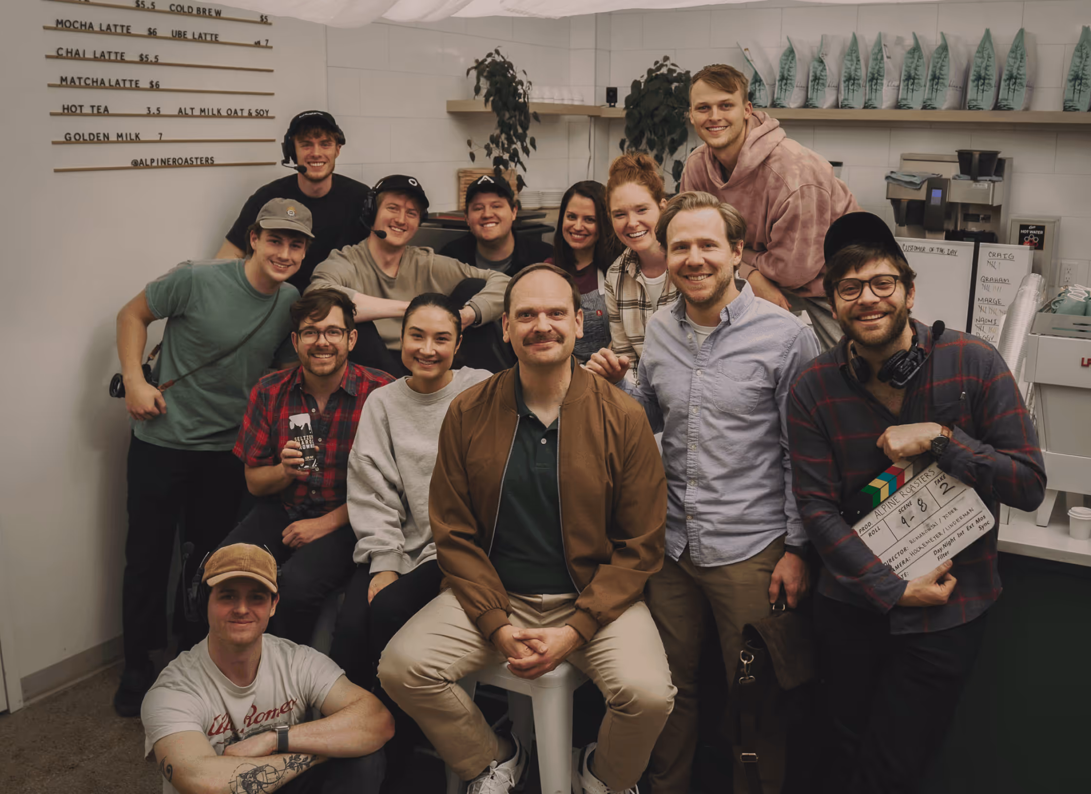 Group of 14 people smiling and posing in a coffee shop with a menu board in the background.