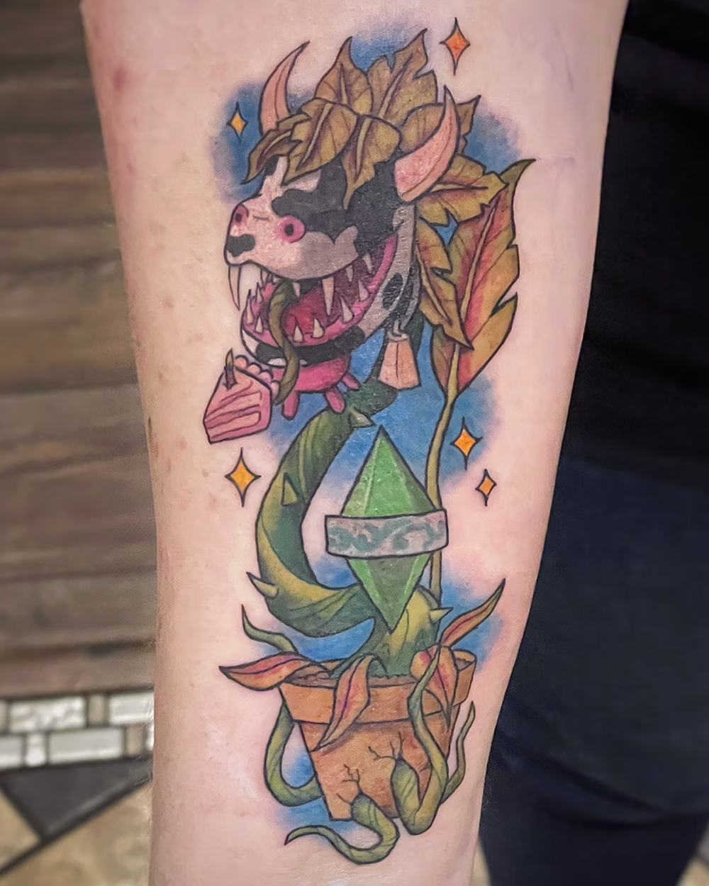 Colorful tattoo of a monstrous plant in a cracked pot with a cow skull and pink tongue, green vines, and autumn leaves.