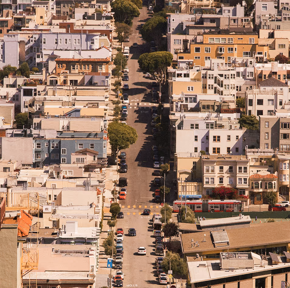 A street lined with parked cars and trees runs between buildings