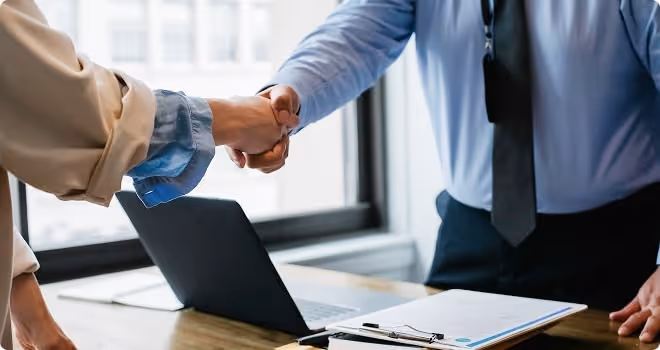 Two people shake hands over a desk with a laptop