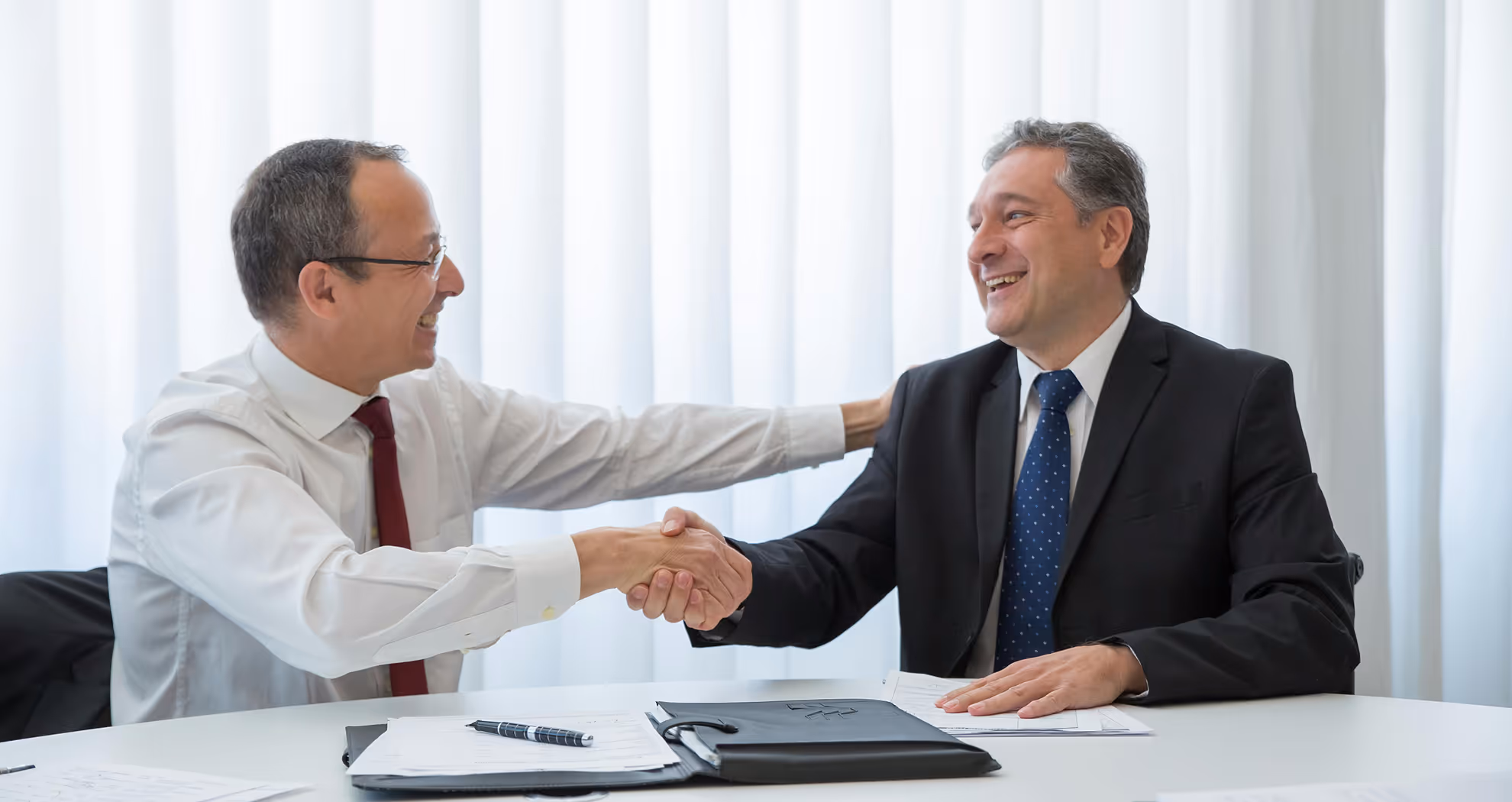 Two smiling businessmen in suits shake hands over a table