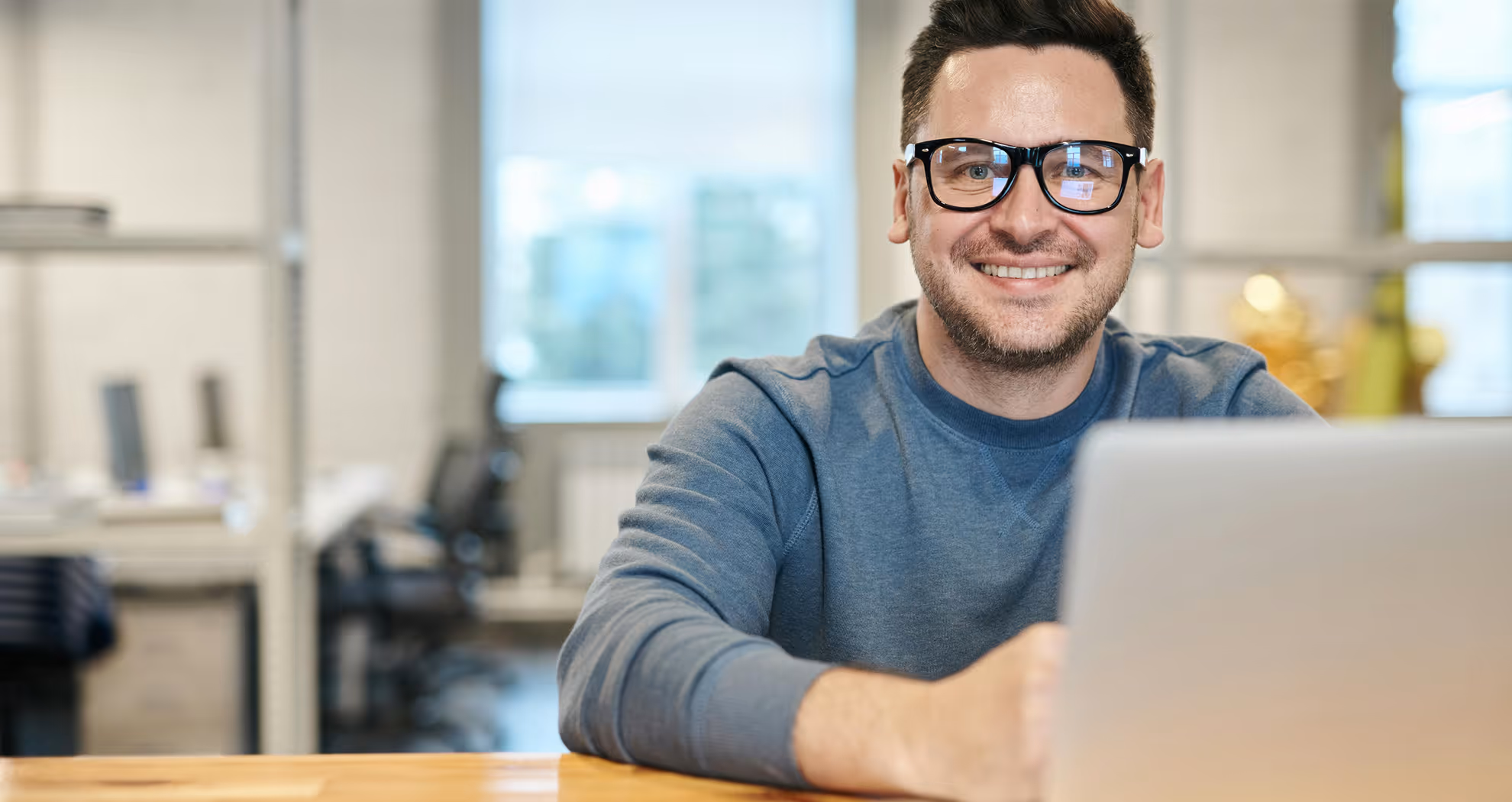 A smiling man with glasses sits at a desk with laptop