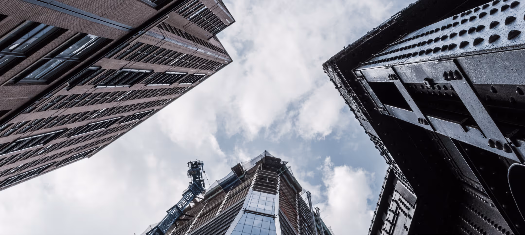 Upward view of three tall buildings against a cloudy sky, including a glass and steel skyscraper under construction with a crane.