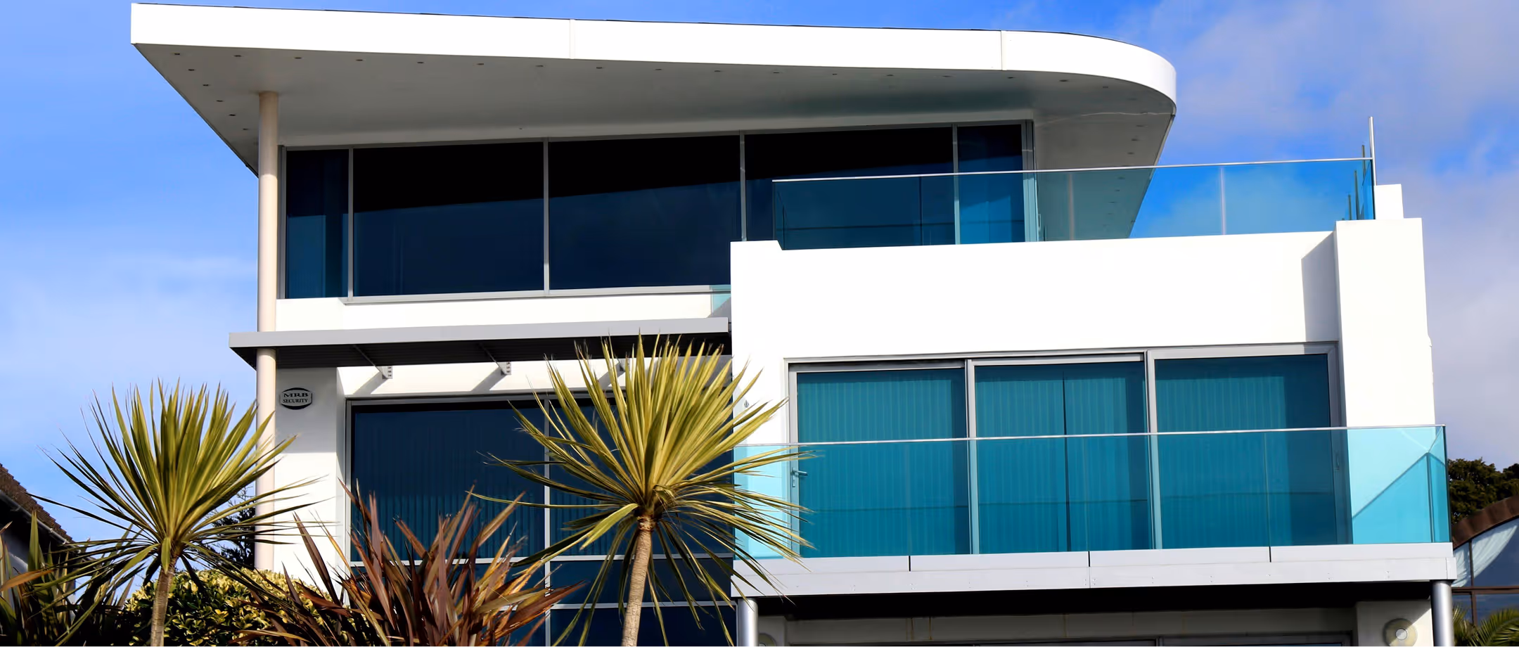 Modern white two-story house with large blue-tinted windows and glass balconies under a blue sky.
