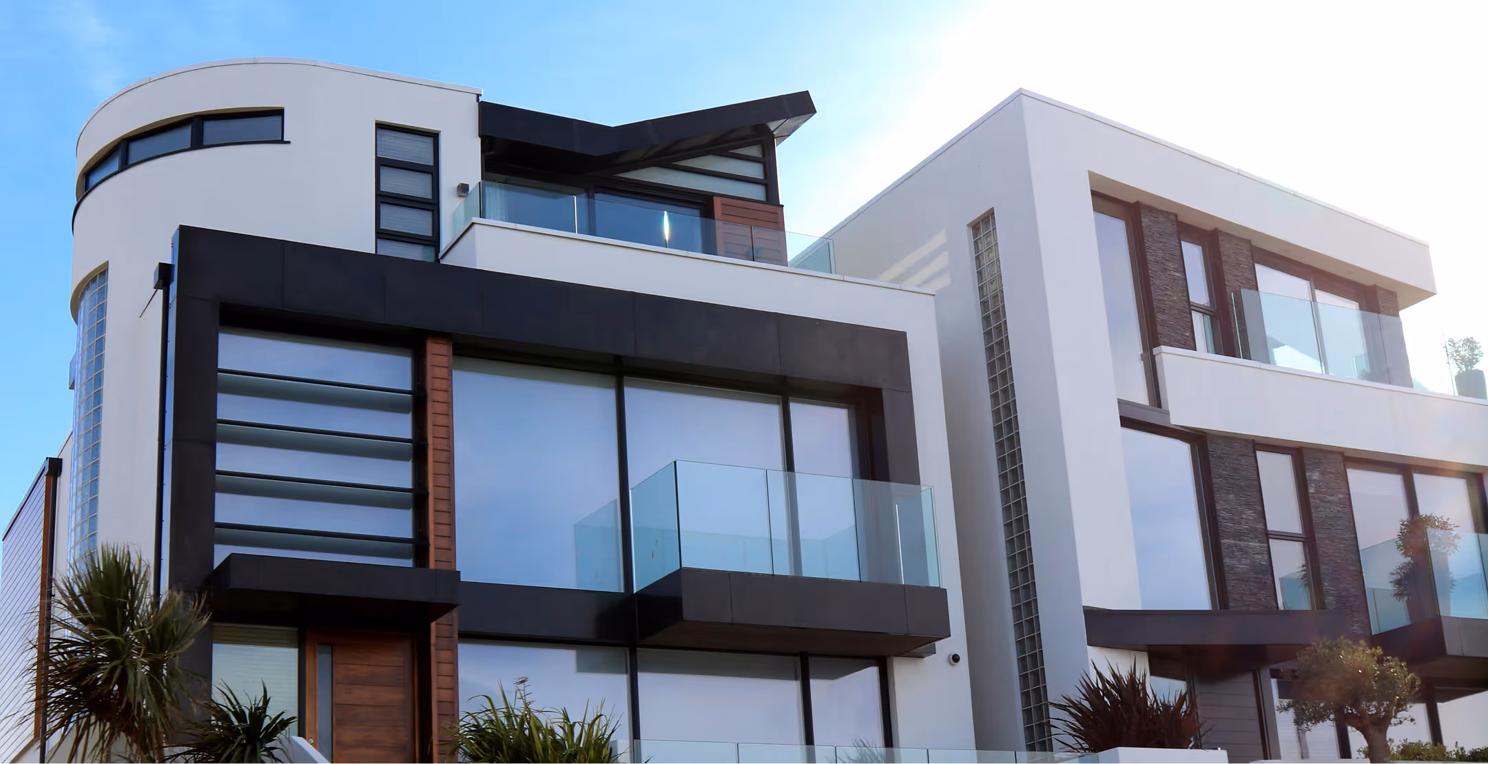 Modern multi-story house with large glass windows, black framing, and balconies with glass railings under a clear blue sky.