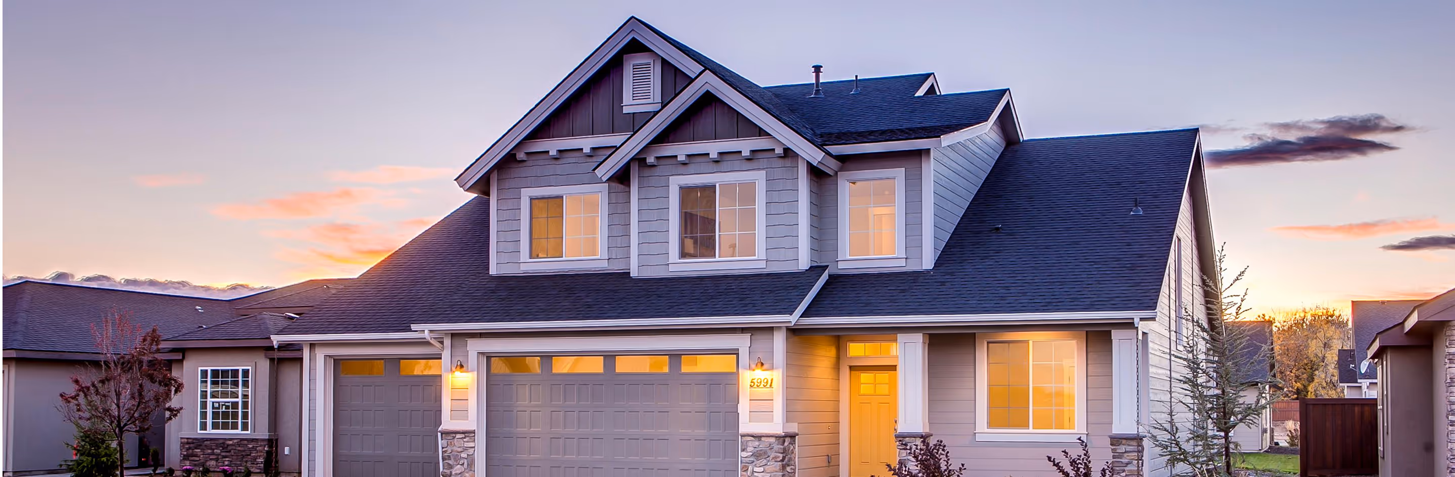 Modern two-story house with gray siding, lit windows, and a double garage at sunset.