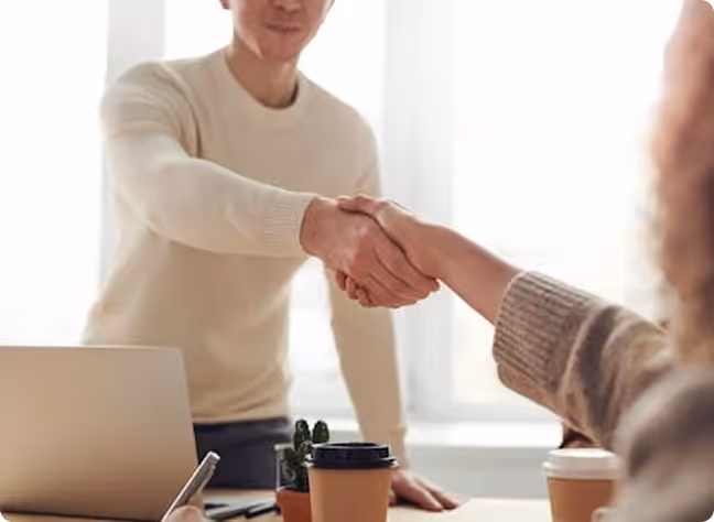 Two people shaking hands over a desk with coffee cups and a laptop in a bright office setting.