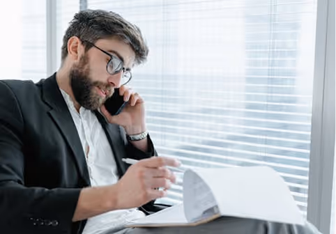 Man with glasses and a beard talking on a phone while reviewing documents in a bright office.