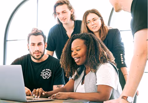 A diverse group of five young adults collaborating and smiling around a laptop in a bright room.