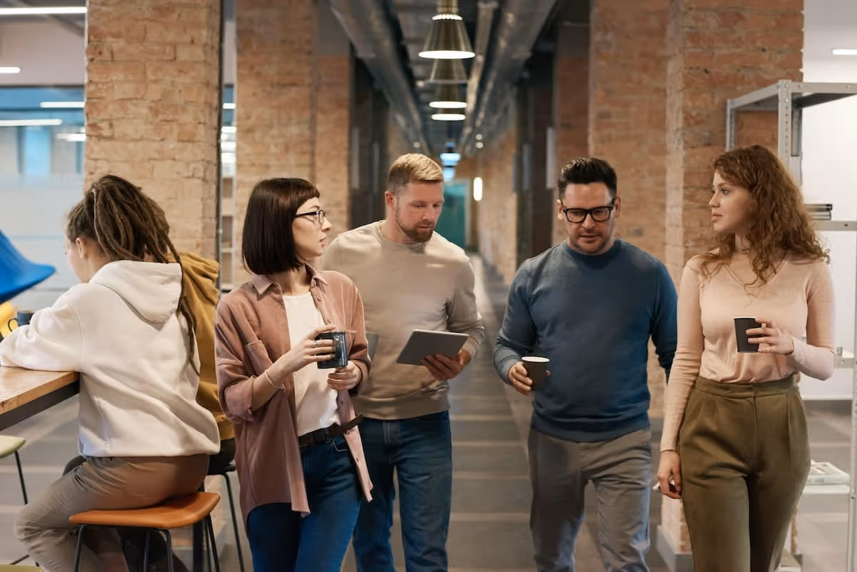 Five colleagues in casual attire conversing and holding coffee cups in a modern office hallway with exposed brick walls.