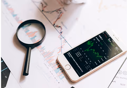 Smartphone displaying a stock market chart with green and red lines, lying on financial documents beside a magnifying glass.