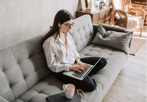 Woman wearing glasses sitting cross-legged on a gray sofa, working on a laptop with a coffee mug and book nearby.