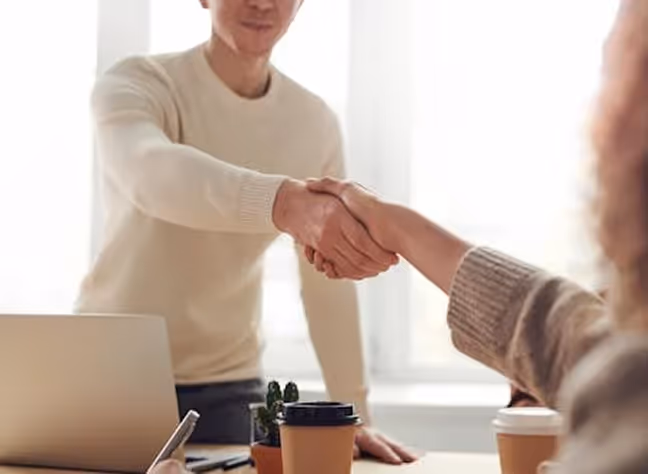 Two people shaking hands over a desk with a laptop, coffee cups, and a small plant.