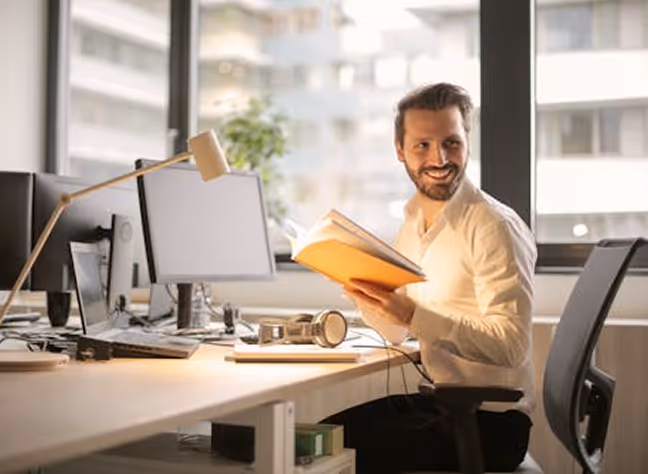 Smiling man holding an open notebook while sitting at a desk with a computer and headphones in a bright office.