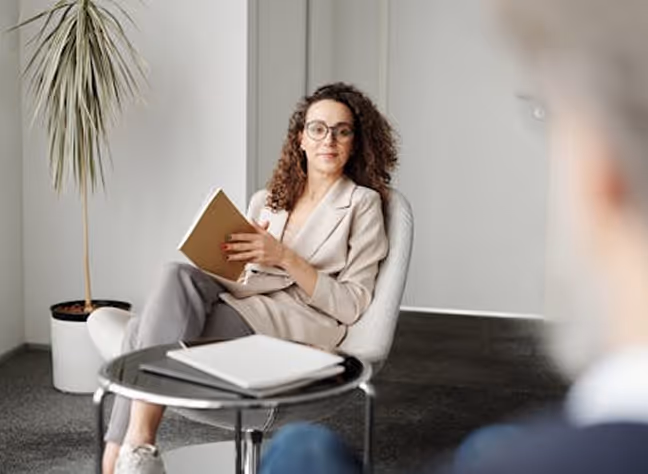 A woman with curly hair and glasses sits in a chair holding a notebook, facing a person whose blurred back is visible.