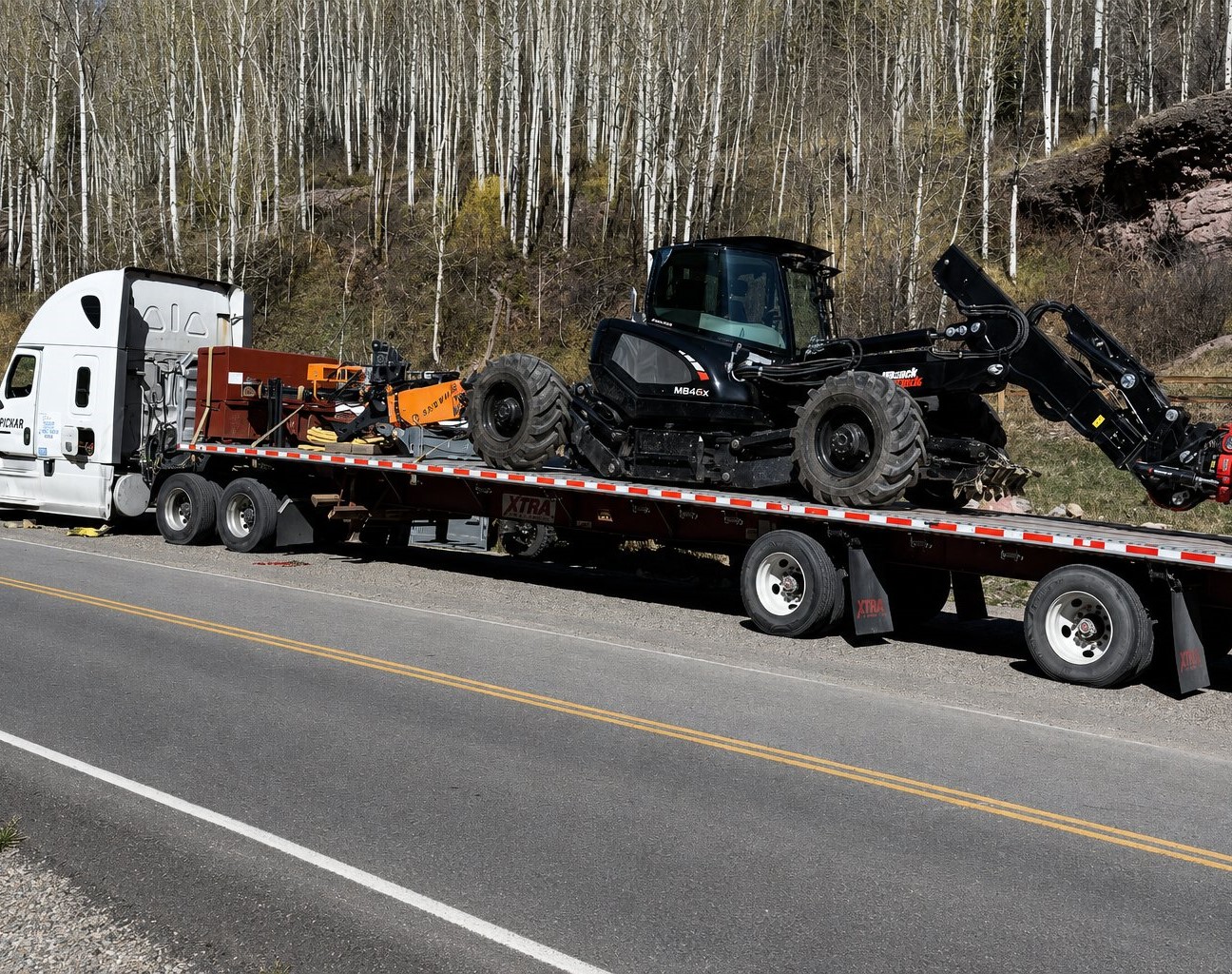 Wheeled excavator secured on a stepdeck flatbed trailer pulled by a semi truck on a mountain road lined with aspen trees