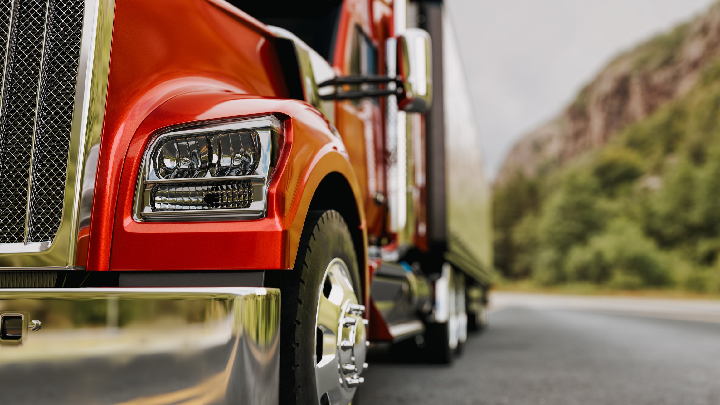 Close low angle view of a red semi truck cab with chrome grille and wheels parked on a highway with hills behind