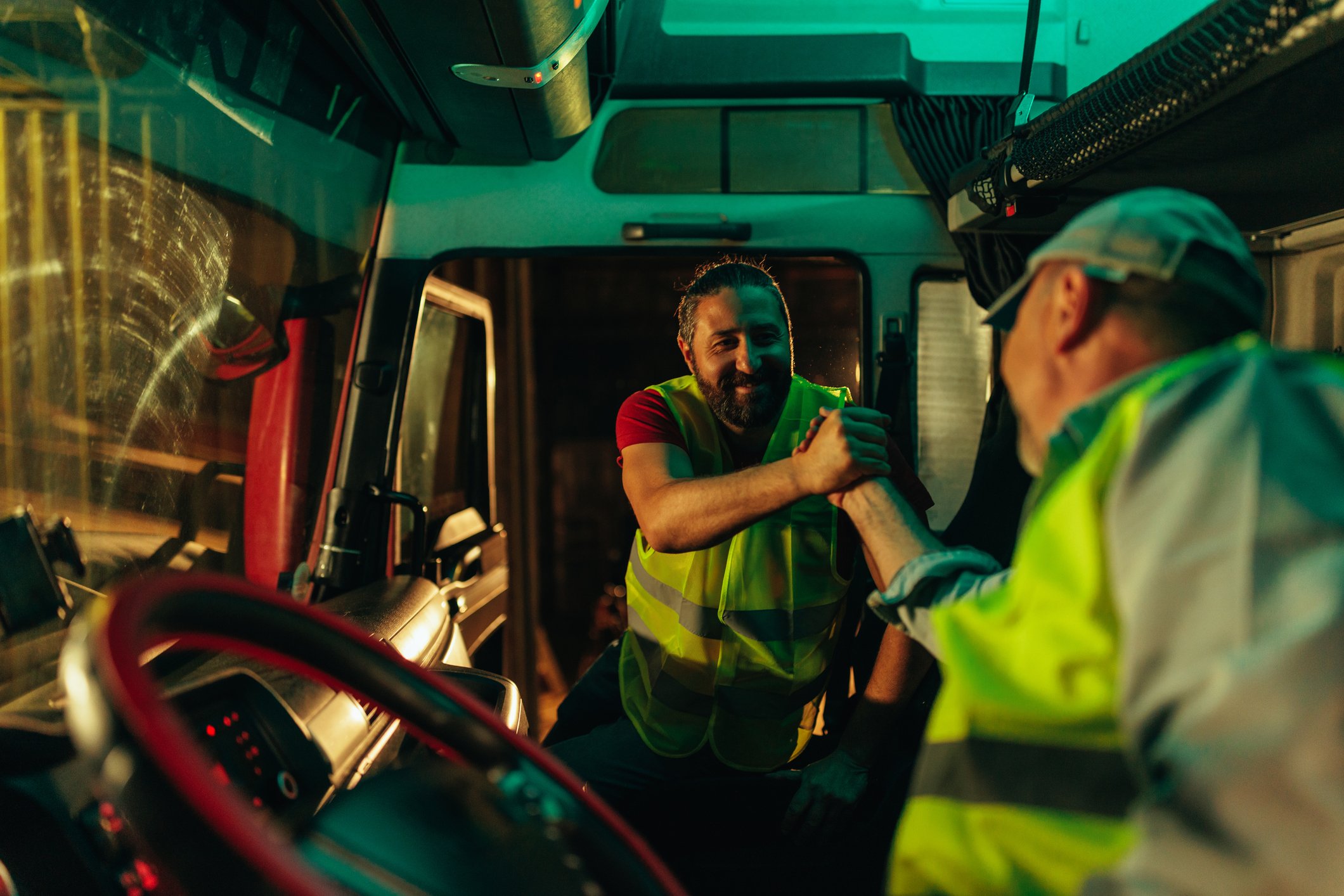 Two truck drivers in high visibility vests clasping hands inside a truck cab during a driver changeover