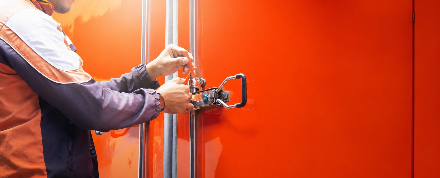 Worker in a safety vest applying a security seal to the door lock rods of an orange trailer