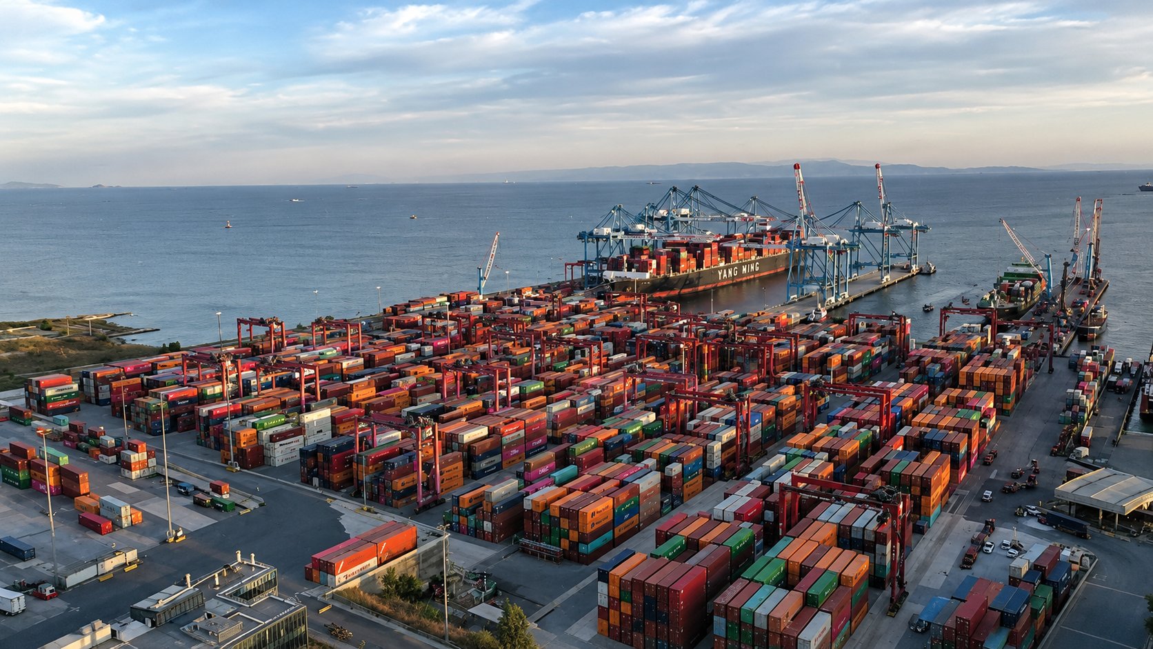 Dense stacks of shipping containers under straddle carriers in a Turkish container terminal yard with a vessel working at berth and the Sea of Marmara behind, an Istanbul area origin port for ocean freight to Los Angeles
