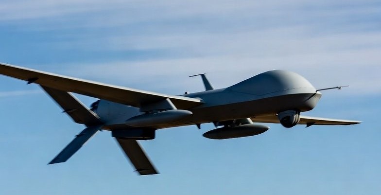 Gray military unmanned aerial vehicle in flight against a blue sky with mountains below