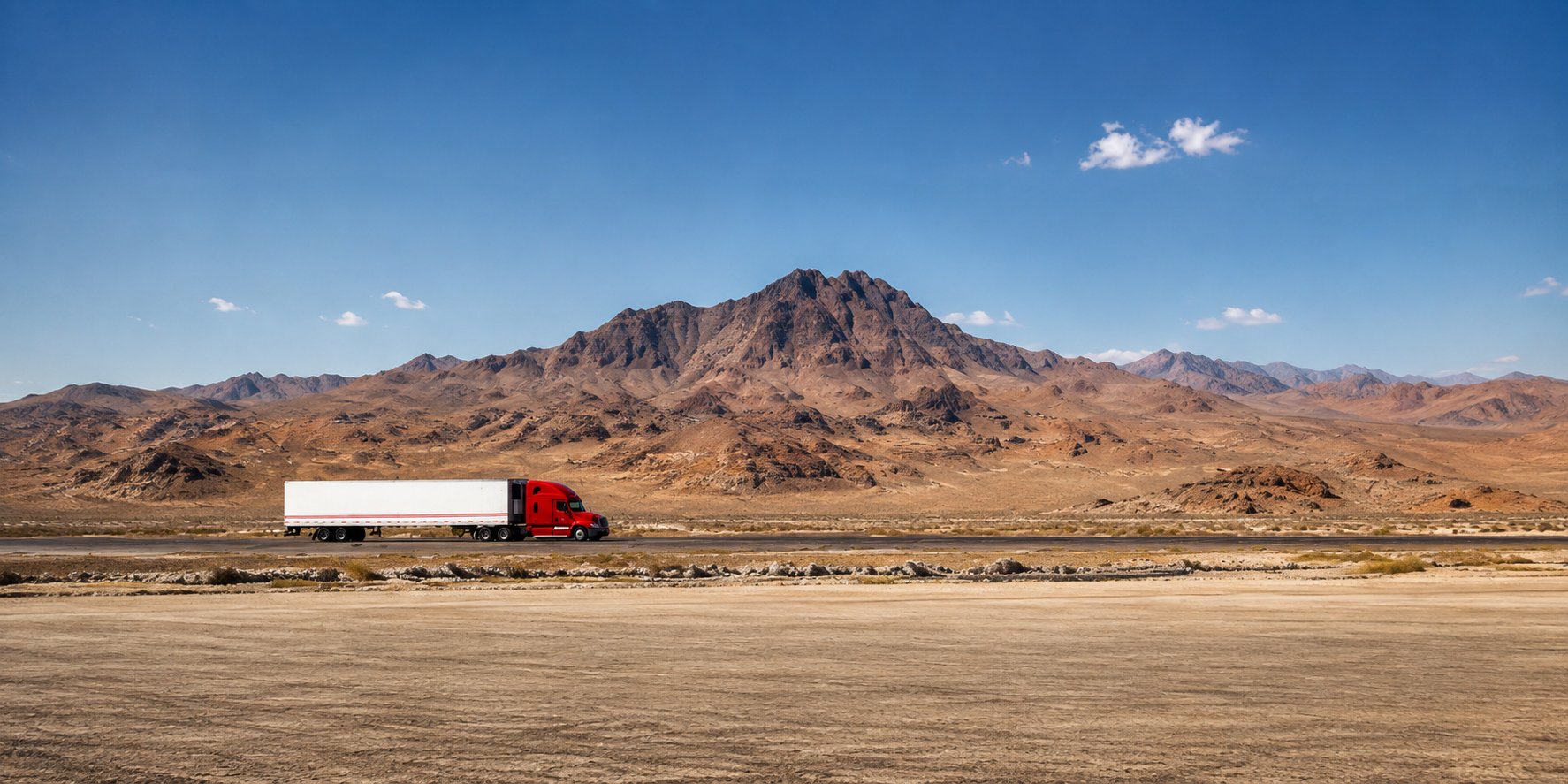 Red semi truck pulling a white dry van trailer across a desert highway with arid mountains behind