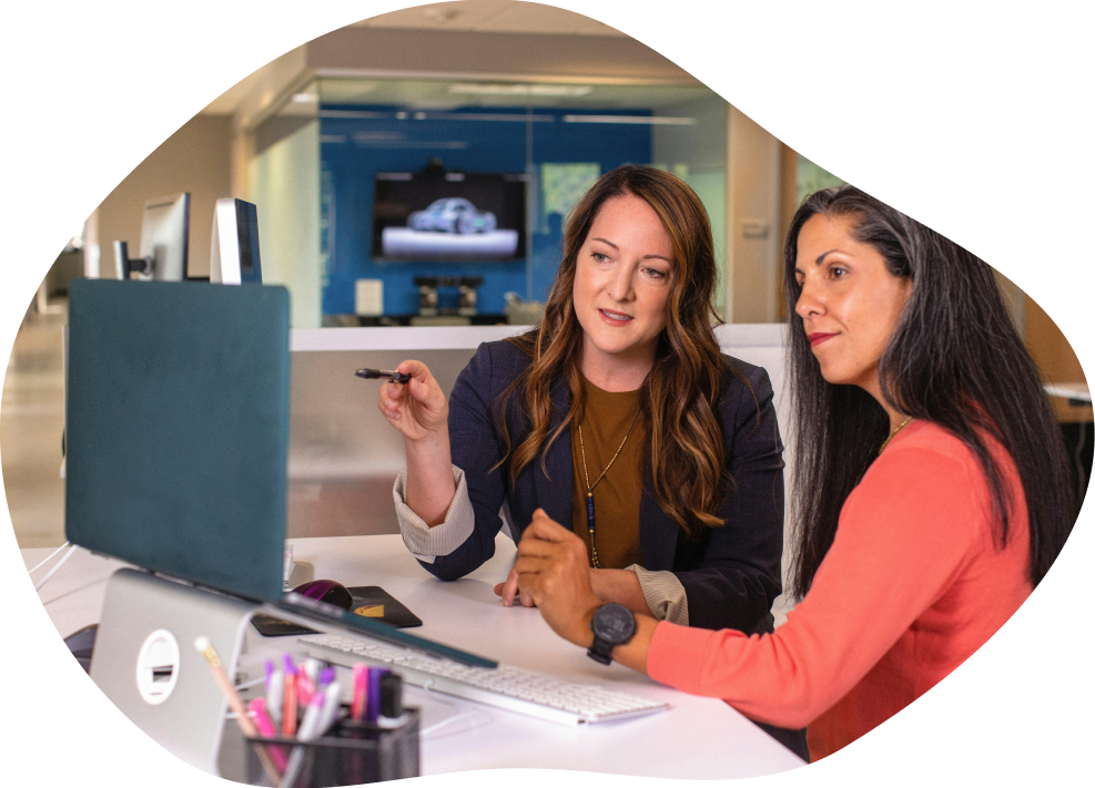 Two women sitting at a desk, one pointing at a computer screen during a discussion in an office.