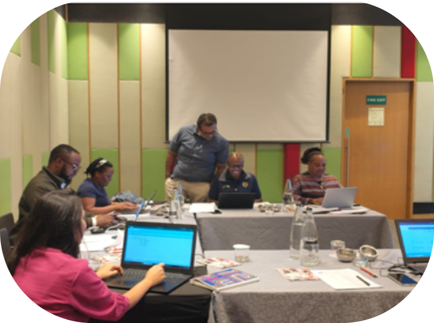 A group of six people working on laptops around a conference table in a meeting room with a blank projector screen and a fire exit door.