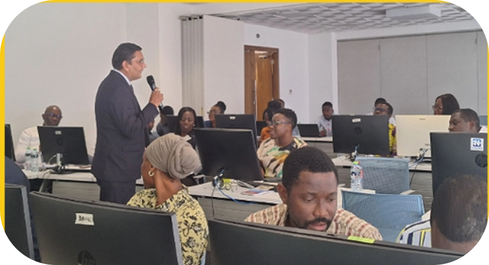 Man in suit speaking into a microphone to a group of people seated at desks with computers in a conference room.