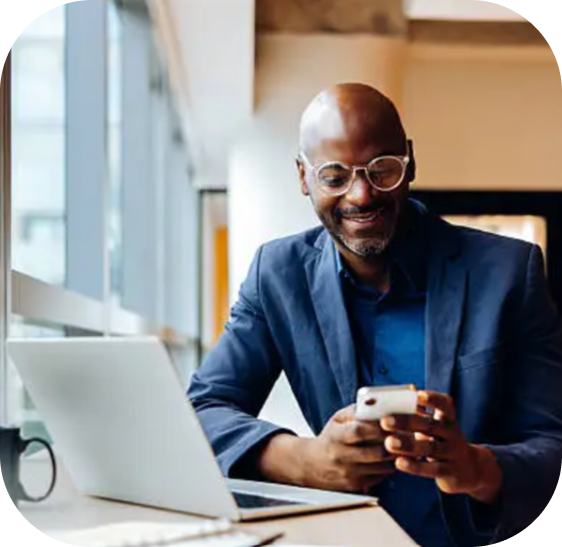 Smiling man in glasses and blazer using smartphone at desk with laptop in a bright office.