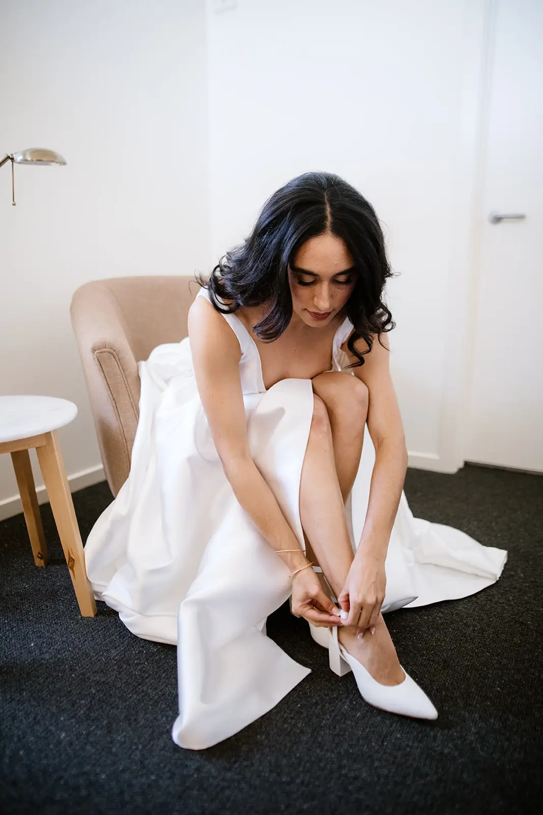 A bride sitting in a chair during her wedding preparations, putting on a white pointed-toe block heel pump. The photo, taken by Brendan Creaser, highlights the thick, stable heel design that is ideal for all-day comfort and navigating outdoor Yarra Valley wedding venues