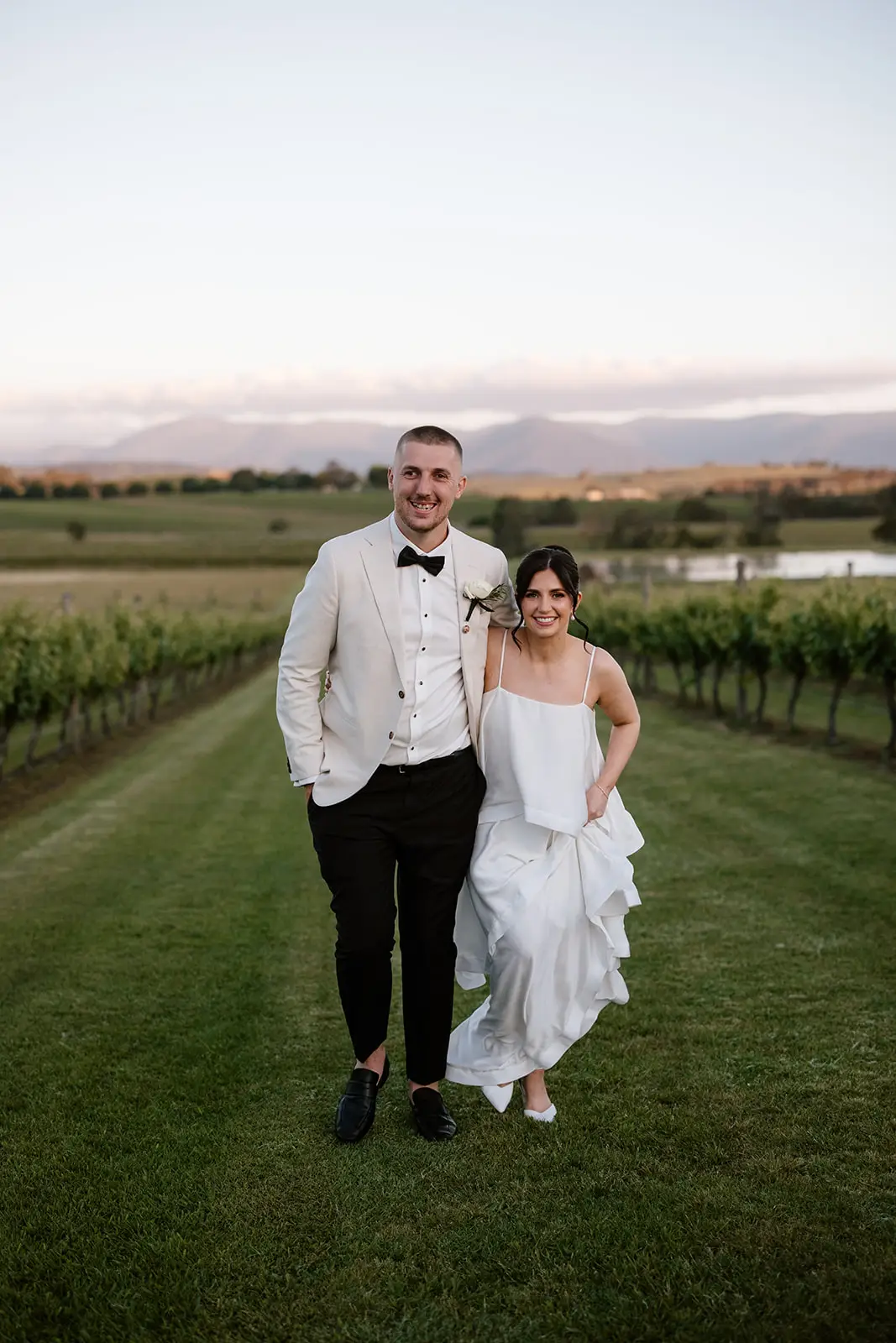 A bride and groom walking through the vineyard rows at sunset. The bride is wearing white kitten heels, demonstrating how a lower, more stable heel height allows for comfortable movement across grass and uneven vineyard terrain without sinking.