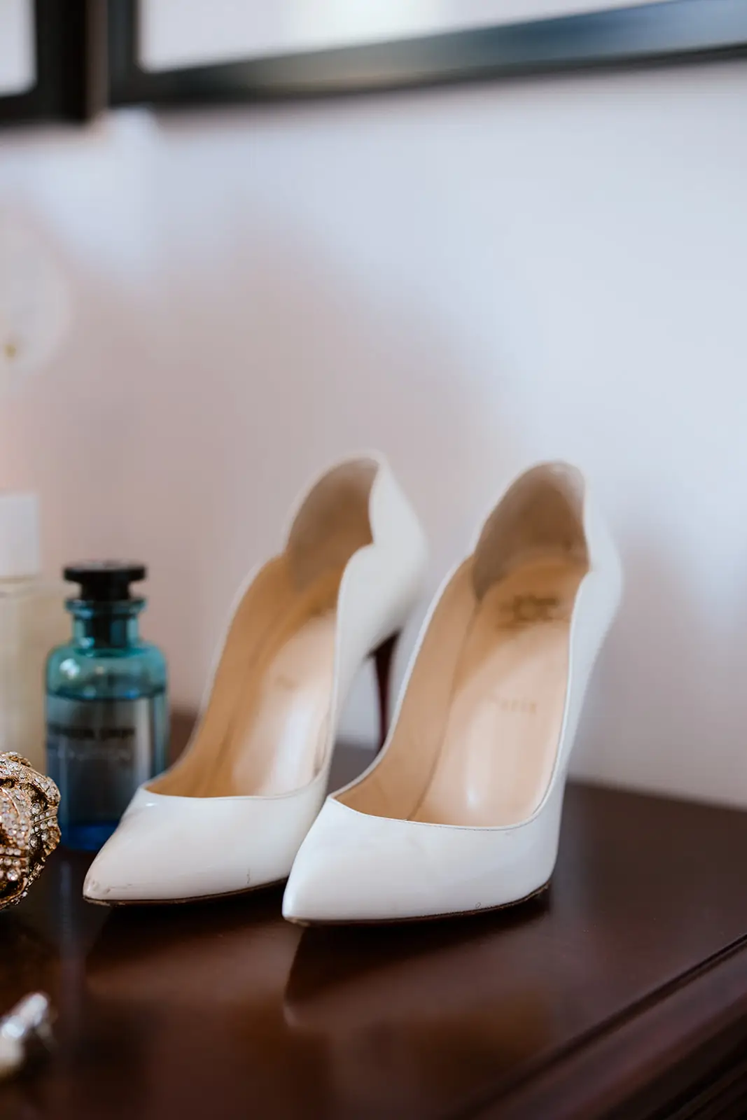 A close-up, soft-focus detail shot of a pair of white Christian Louboutin "Hot Chick" pointed-toe stiletto pumps sitting on a dark wooden dresser. The shoes feature a distinct scalloped edge along the opening. In the background, a blue glass perfume bottle and a sparkling gold accessory are visible under warm, indoor light.