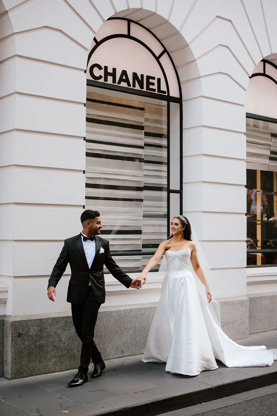 A bride and groom walking past the Chanel boutique storefront in Melbourne. The bride is wearing a classic white ballgown and white heels, perfectly illustrating the polished city wedding aesthetic discussed in this guide. Photo captured by Brendan Creaser.